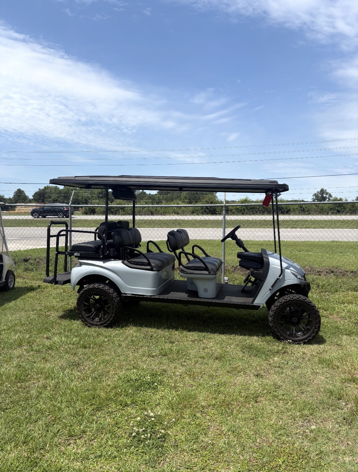 Light gray six-passenger lifted electric golf cart with black seats, black wheels, and a rear flip seat parked on grass outdoors.