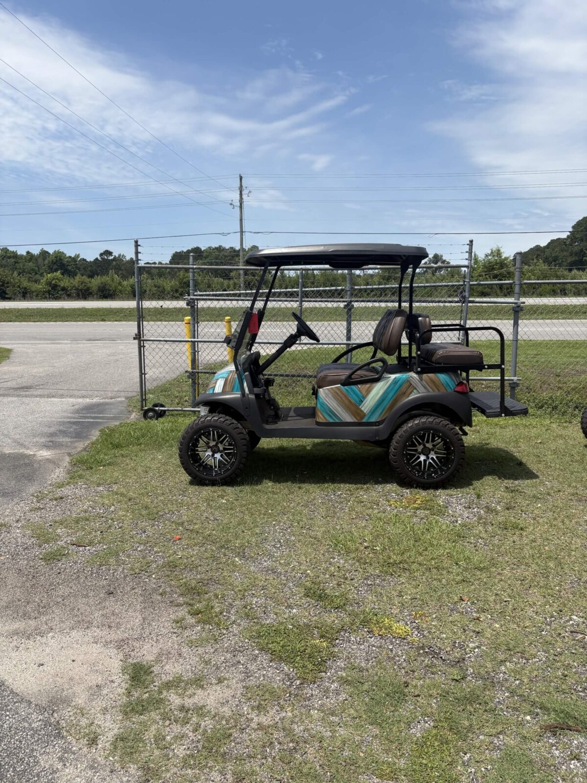 2022 Club Car Precedent gas golf cart with custom turquoise and woodgrain body panels, lifted suspension, black wheels, and rear flip seat parked outdoors.